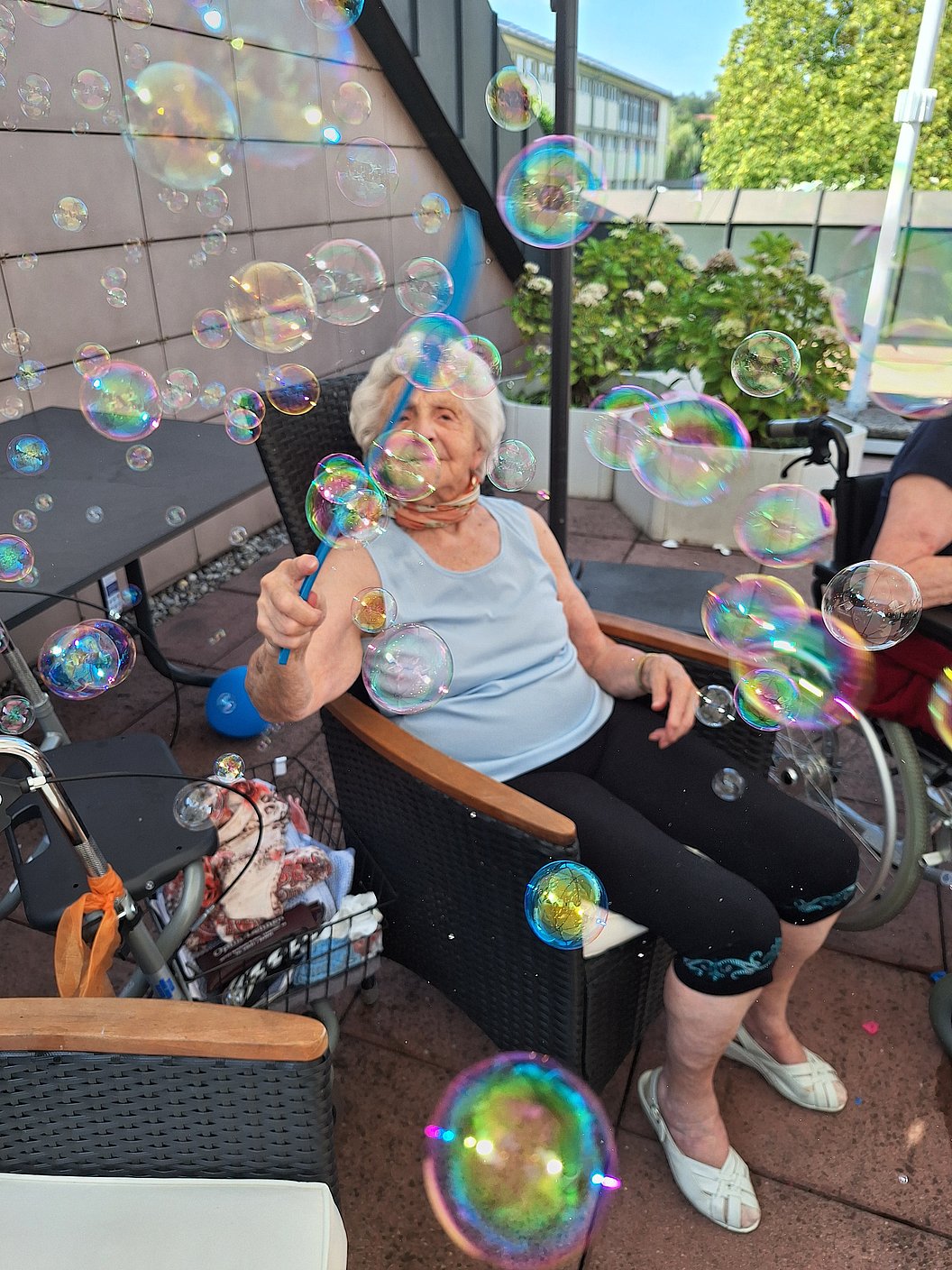 Ältere Person sitzt auf Terrasse in schwarzem Korbsessel, umgeben von vielen schillernden Seifenblasen.