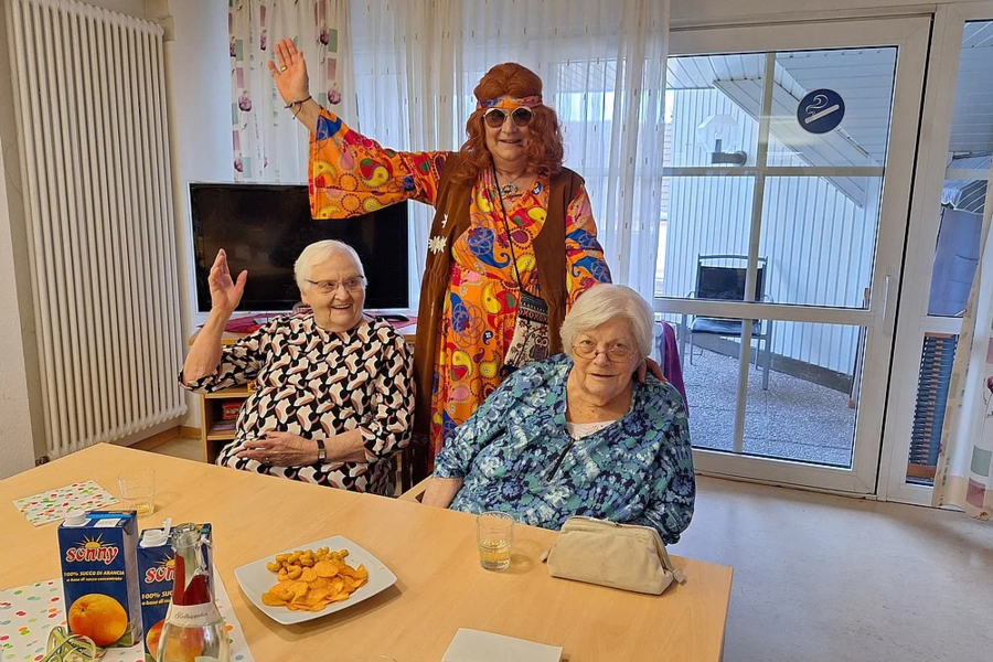 Drei Personen in einem hellen Raum, zwei sitzen am Tisch mit Snacks und Getränken, eine steht dahinter mit erhobenen Armen und bunter Kleidung.