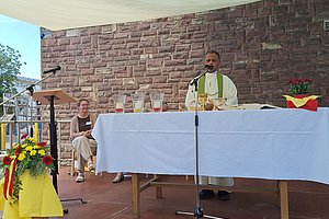 Priester steht hinter einem mit weißem Tuch bedeckten Altar auf einer Bühne vor einer roten Backsteinwand, links Blumenarrangement und Mikrofonständer, im Hintergrund zwei sitzende Personen