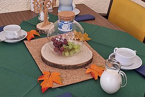 Tisch mit grünem Tischtuch, darauf Holzscheibe mit Trauben, Wasserflasche, Milchkännchen, Kaffeetassen und herbstliche Dekoration mit Blättern und Baumfigur