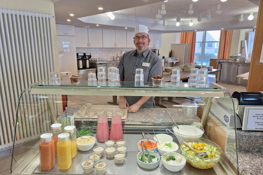Mann in grauem Hemd steht hinter einer Glasvitrine mit verschiedenen Smoothies, Schalen mit Gemüse und Salat, sowie kleinen Schälchen mit Zutaten.