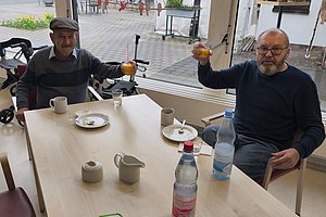 Zwei Personen sitzen an einem Tisch mit Getränken und Tellern, eine Person hält ein Glas in die Luft, im Hintergrund Fenster mit Blick auf einen Innenhof