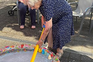 Ältere Frau mit blau-weiß gepunktetem Kleid beugt sich über ein kleines Planschbecken und hält einen gelben Gegenstand, im Hintergrund sitzen zwei ältere Frauen auf Stühlen.