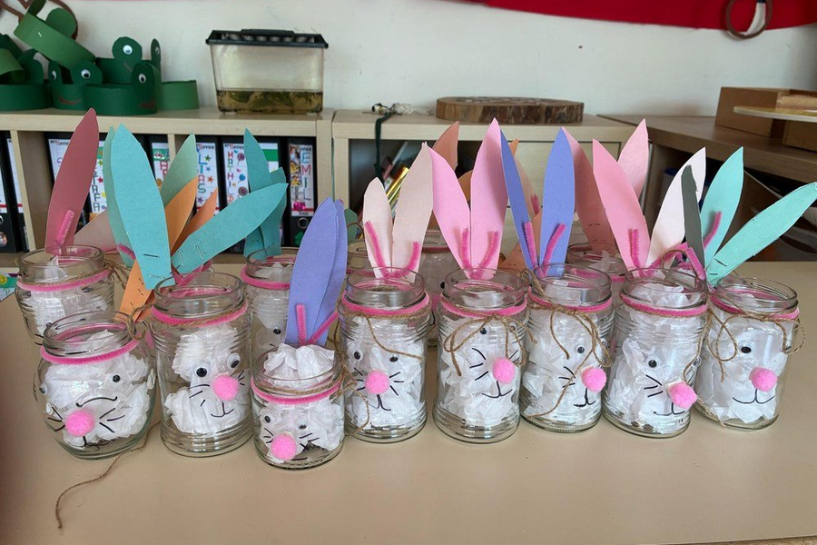 Mehrere Einmachgläser mit weißen Hasengesichtern aus Watte, rosa Nasen und bunten Papierohren in Blau, Rosa und Lila auf einem Tisch.
