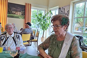 Mehrere ältere Personen sitzen an einem Tisch in einem hellen Raum mit großen Fenstern und sichtbaren Holzbalken im Dach.