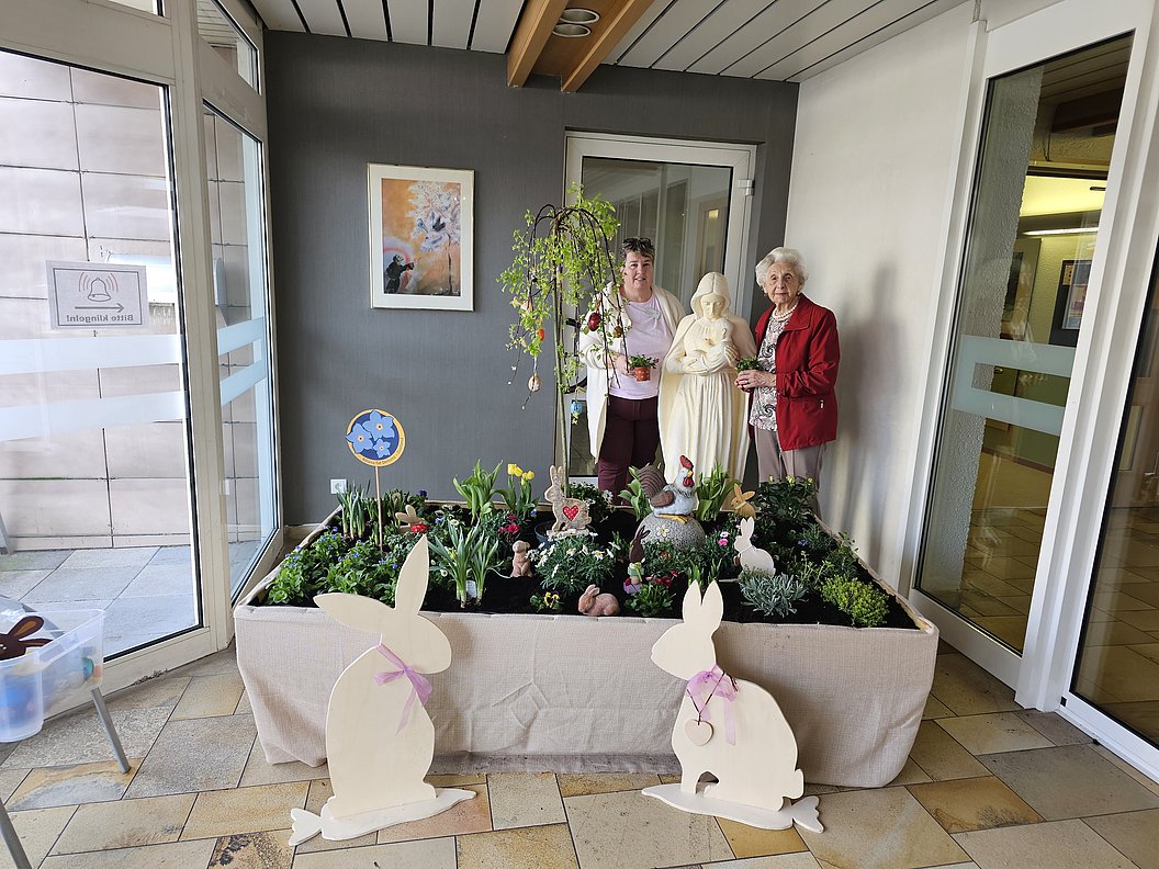 Zwei Personen stehen neben einer Statue in einem rechteckigen Pflanzbeet mit grünen Pflanzen und zwei weißen Hasenfiguren mit rosa Schleifen davor.
