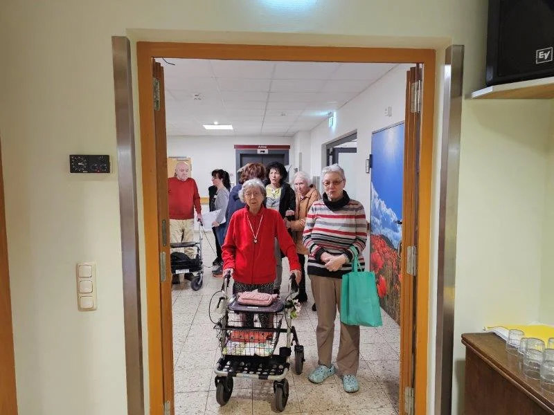 Mehrere ältere Menschen in einem Flur, eine Frau mit Rollator und eine andere mit grünem Einkaufstaschenbeutel, weitere Personen im Hintergrund.
