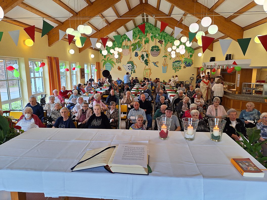 Großer Festsaal mit vielen sitzenden älteren Menschen, bunte Wimpelketten und hängende Pflanzen an der Decke, vorne ein mit weißem Tuch bedeckter Tisch mit offenem Buch und Kerzen.