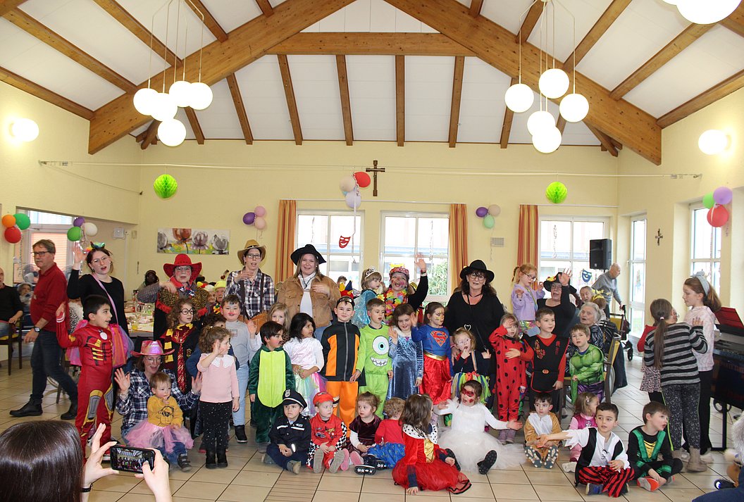 Gruppe von Kindern und Erwachsenen in bunten Kostümen in einem Saal mit Holzbalkendecke und Lampen, bunte Luftballons an den Wänden.