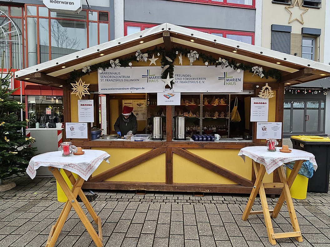 Gelber Holzstand mit Weihnachtsdekoration, zwei Stehtische mit weißen Tüchern davor, Person mit roter Mütze hinter dem Tresen.