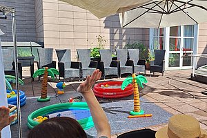Mehrere Personen sitzen im Freien auf Stühlen unter Sonnenschirmen neben aufblasbaren Planschbecken und Spielzeugpalmen auf einem gepflasterten Hof.