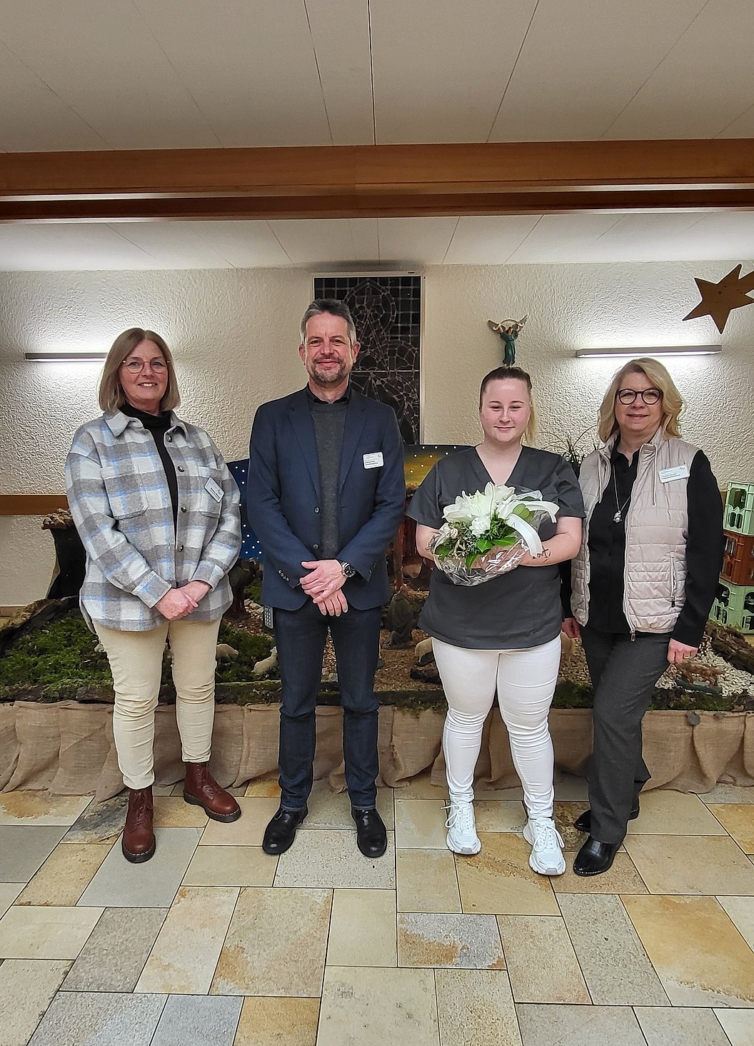 Vier Personen stehen nebeneinander in einem Raum mit Steinboden und Wanddeko, eine Person hält einen Blumenstrauß.