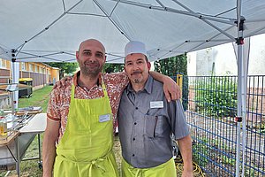 Zwei Männer mit grünen Schürzen stehen unter einem weißen Pavillon und lächeln in die Kamera