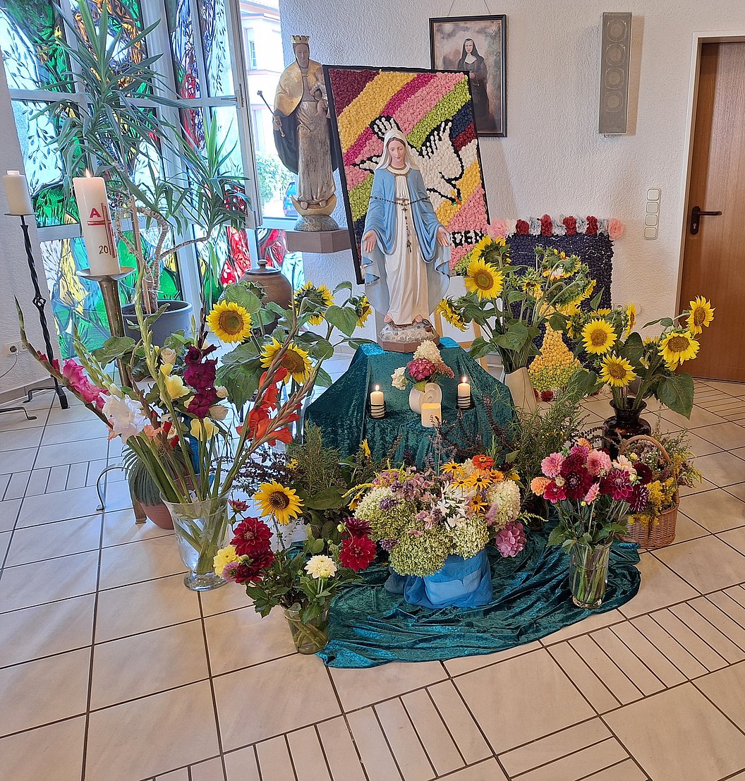 Altar mit Marienstatue, umgeben von Sonnenblumen, weiteren Blumensträußen, Kerzen und religiösen Bildern in einem Raum mit Fliesenboden