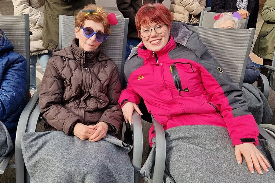 Zwei Personen sitzen nebeneinander auf grauen Stühlen, beide mit grauen Decken bedeckt, eine trägt eine pinke Jacke, die andere eine braune Jacke.