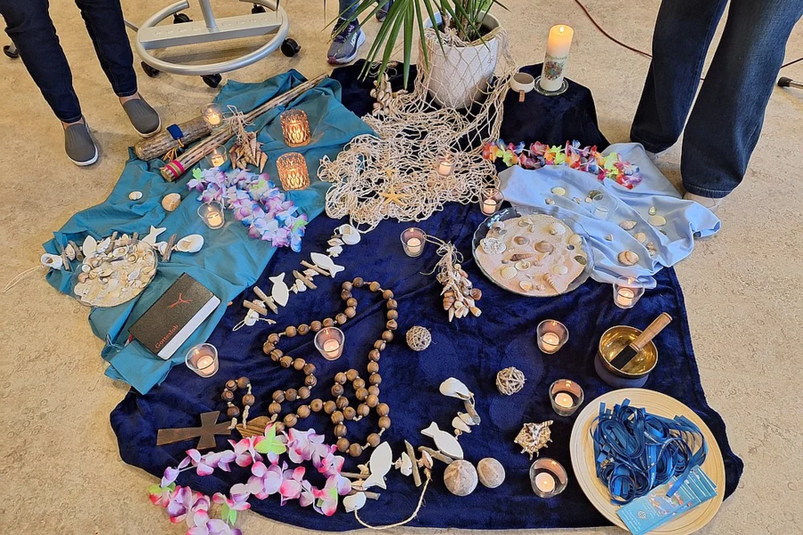 Mehrere bunte Tücher mit Muscheln, Kerzen, Holzperlenketten, Blumenketten und weiteren Dekorationsgegenständen auf dem Boden, umgeben von Personenbeinen.