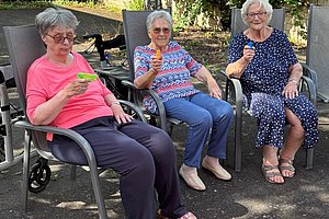 Drei ältere Frauen sitzen auf Gartenstühlen im Freien und halten jeweils ein Getränk in der Hand