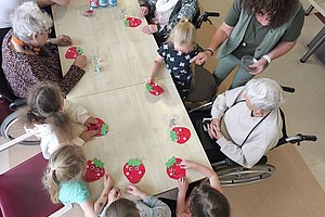 Mehrgenerationentisch mit Kindern und älteren Menschen, die an Bastelarbeiten mit roten Erdbeerformen sitzen