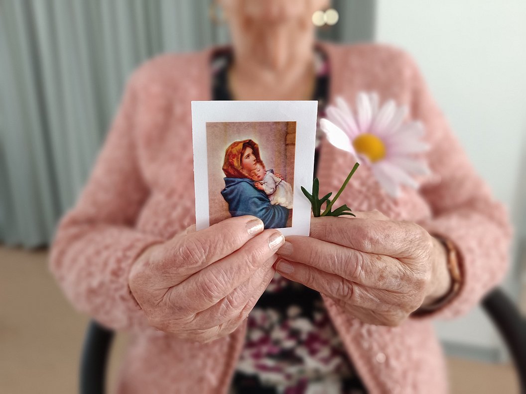 Ältere Person hält ein Foto eines Kindes mit Kopftuch und eine einzelne Blüte mit weißen Blütenblättern und gelber Mitte.