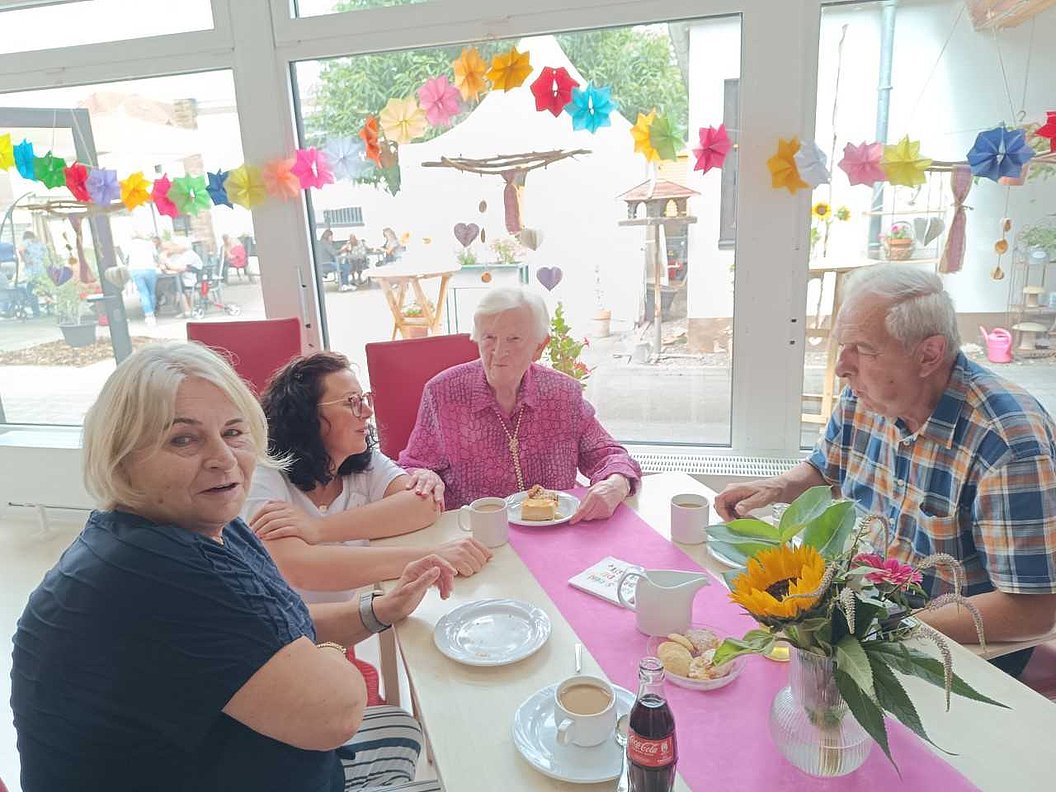 Vier Personen sitzen an einem Tisch mit rosa Tischdecke, darauf Tassen, Teller, eine Flasche Cola und eine Vase mit Sonnenblume.