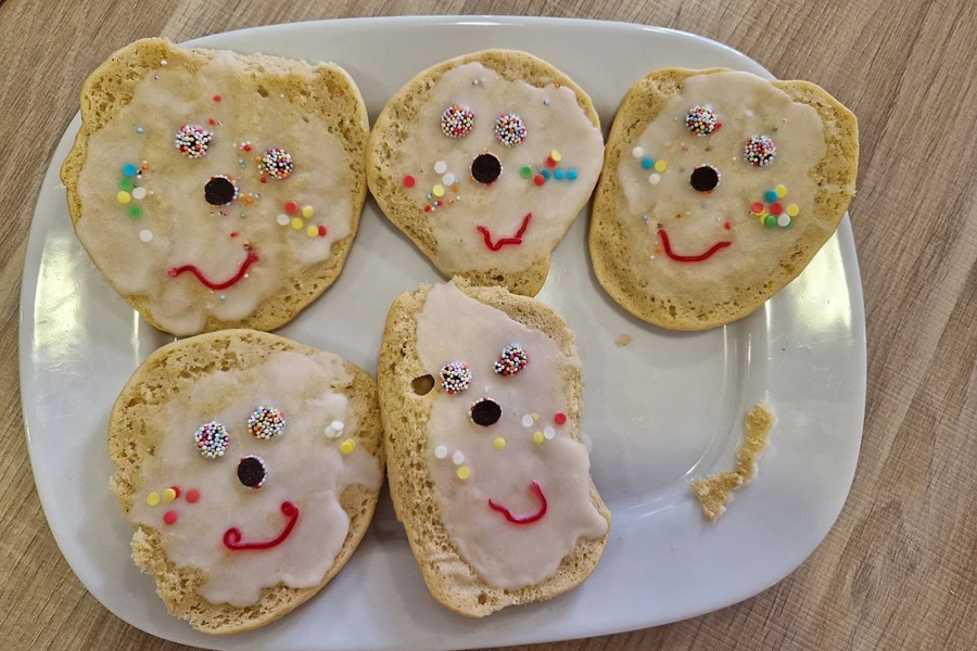 Fünf mit Zuckerguss und bunten Streuseln verzierte Kekse auf einem weißen Teller, die Gesichter mit Augen und lachenden Mündern zeigen.