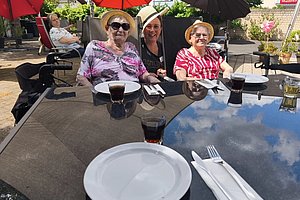 Drei ältere Frauen sitzen an einem Tisch im Freien unter Sonnenschirmen, auf dem Tisch stehen Gläser mit dunklem Getränk und weiße Teller mit Besteck.