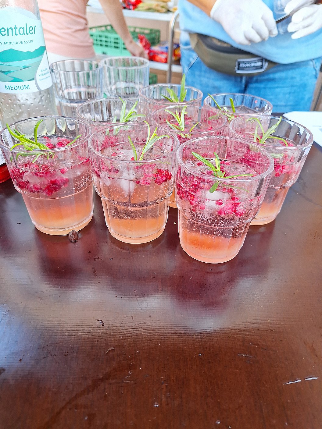 Mehrere durchsichtige Gläser mit rosa Getränk, roten Beeren und grünen Kräutern auf dunklem Tisch.