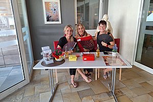 Drei Frauen sitzen an einem Tisch in einem hellen Raum und halten Fächer in der Hand