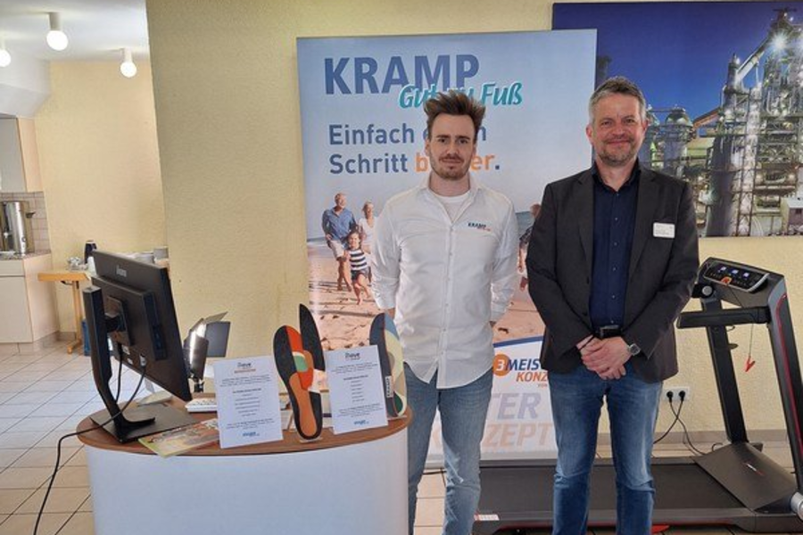Zwei Männer stehen nebeneinander vor einem Messestand mit dem Schriftzug 'KRAMP Gut zu Fuß' auf dem Tresen und einem Plakat im Hintergrund.