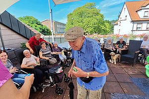 Älterer Mann steht auf einer Terrasse unter Sonnenschirmen, umgeben von sitzenden Menschen und Rollstühlen, bei sonnigem Wetter