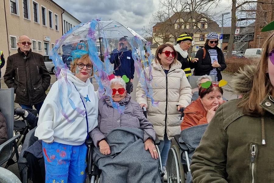 Gruppe von Menschen im Freien, darunter zwei Personen im Rollstuhl, eine mit transparentem Regenschirm mit bunten Mustern.