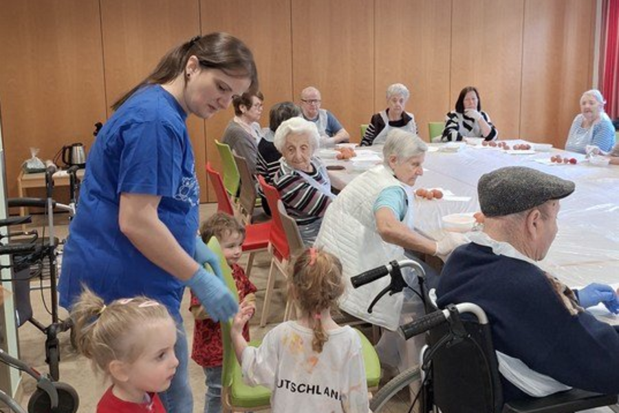 Pflegekraft in blauer Kleidung steht neben mehreren älteren Menschen, einige im Rollstuhl, in einem Raum mit Holzwand und Deckenleuchten.