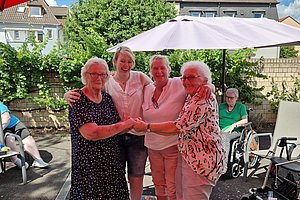 Vier ältere Frauen stehen in einem kleinen aufblasbaren Pool unter einem roten Sonnenschirm und halten sich an den Händen