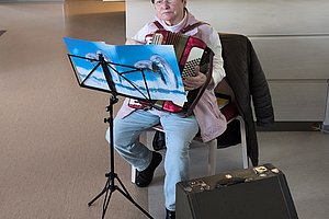 Ältere Frau mit einer Harmonika, sitzt auf einem Stuhl und spielt Musik in einem hellen Raum.