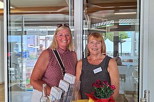 Zwei lächelnde Frauen stehen an einem Stehtisch mit Flyern, einer Flasche und einem roten Blumenstrauß in einem hellen Raum mit Glaswänden