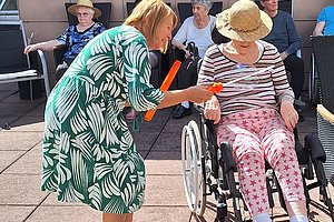 Frau im grünen Kleid mit Blattmuster überreicht einer Frau im Rollstuhl mit Strohhut und gestreiftem Oberteil ein orangefarbenes Objekt
