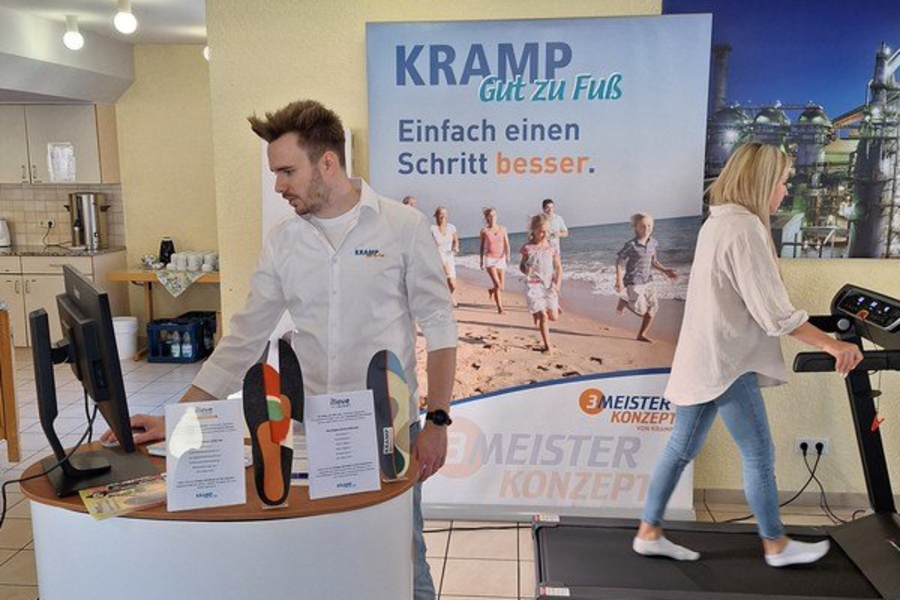 Mann an einem Messestand mit Produkten, im Hintergrund Frau auf einem Laufband, Werbebanner mit Text 'KRAMP Gut zu Fuß Einfach einen Schritt besser.'