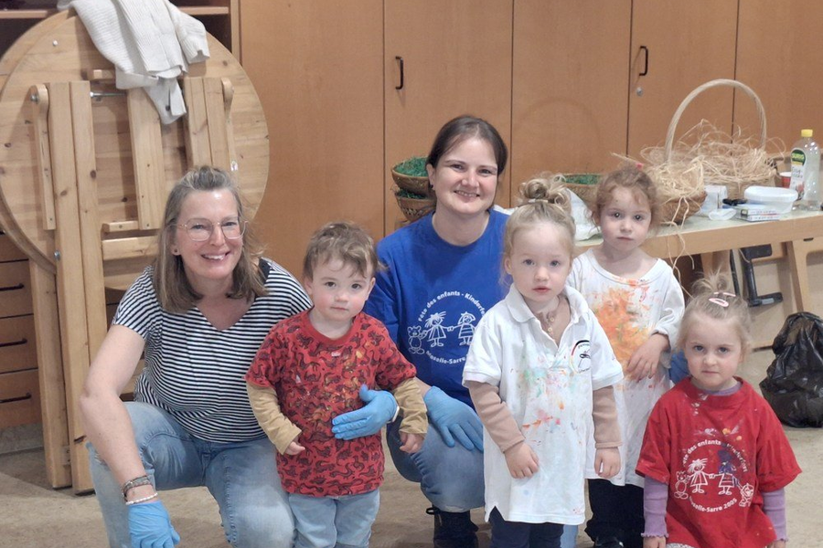 Fünf Kinder und zwei Erwachsene in einem Raum mit Holzmöbeln, die Kinder tragen bunte T-Shirts, die Erwachsenen blaue Handschuhe.