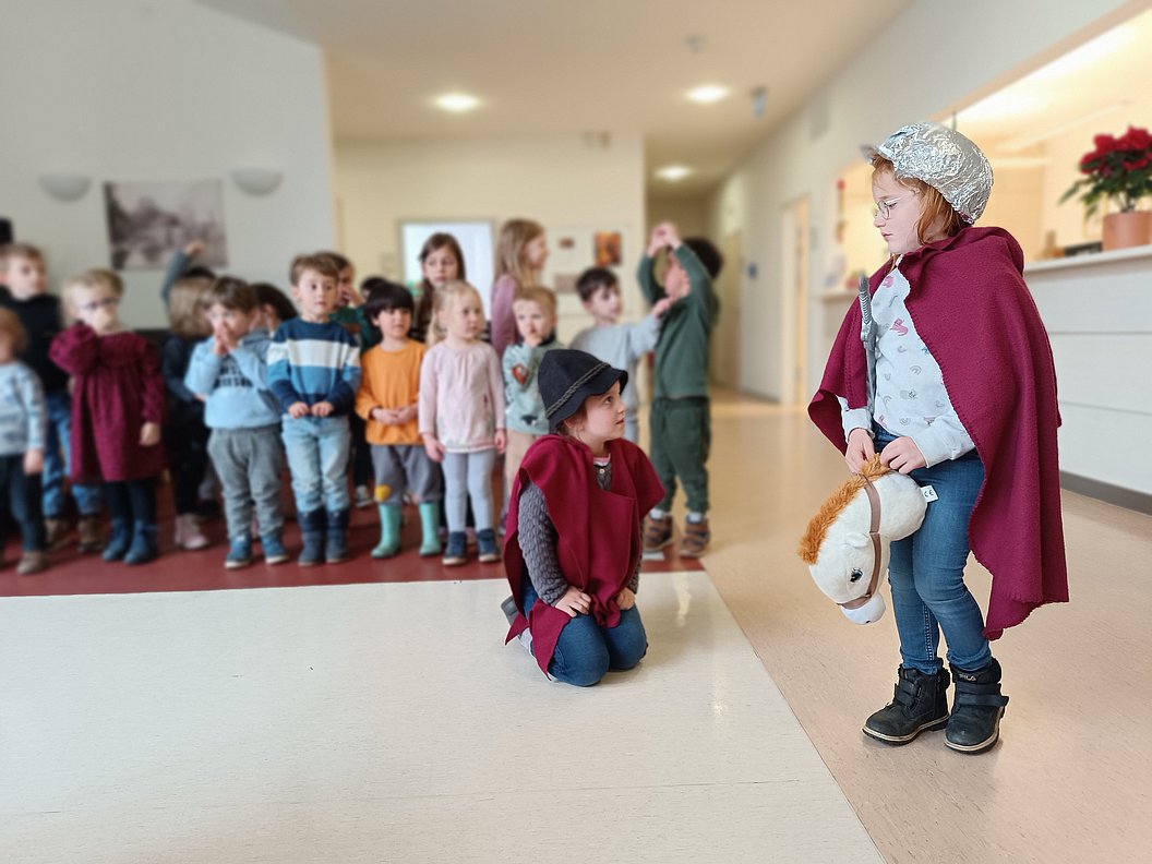 Kind mit Umhang und Haube hält ein Steckenpferd, weitere Kinder stehen und sitzen im Hintergrund in einem Flur.