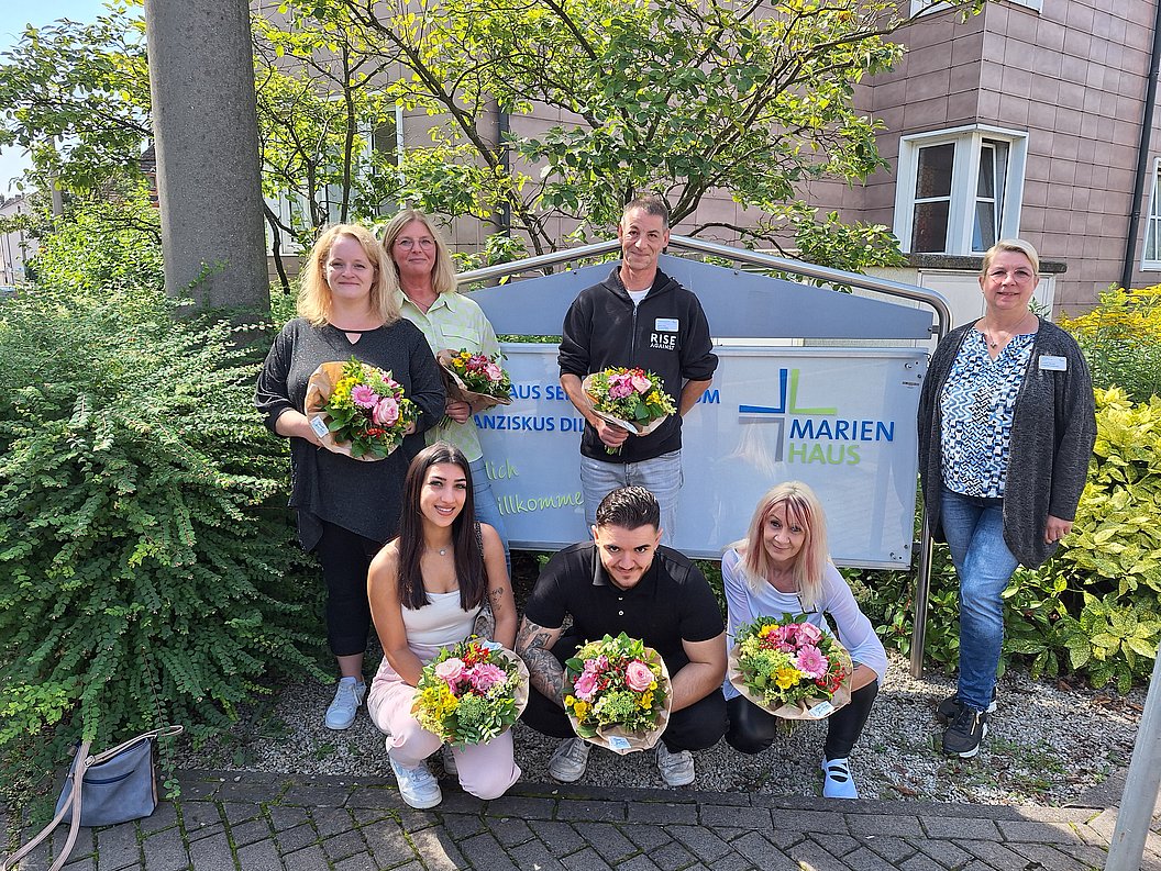 Gruppe von sieben Personen vor einem Schild mit der Aufschrift 'Marien Haus', jede Person hält einen Blumenstrauß.