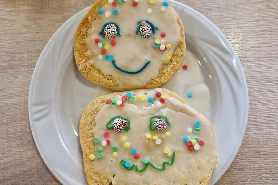 Zwei mit Zuckerguss und bunten Streuseln verzierte Kekse auf weißem Teller, jeweils mit einem lächelnden Gesicht aus Zuckerguss.