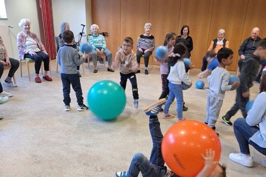 Kinder spielen mit bunten Bällen auf dem Boden, während ältere Menschen auf Stühlen im Hintergrund sitzen