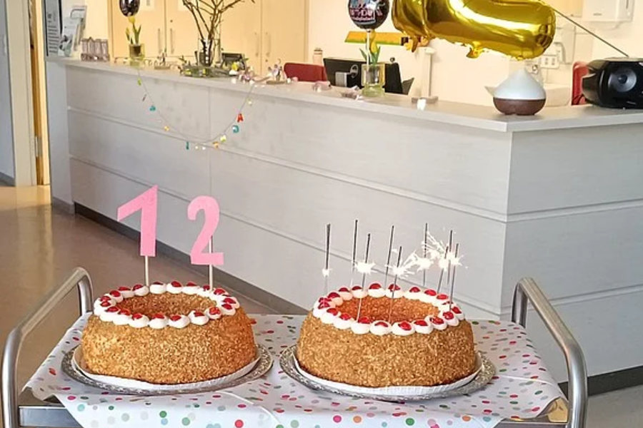 Zwei runde Kuchen mit Kerzen und rosa Zahlenkerzen '1' und '2' auf einem Servierwagen vor einer Theke mit goldener Ballonzahl '2' und Geburtstagsgirlande.