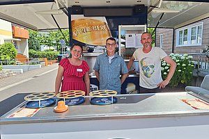Drei Personen stehen hinter einem Verkaufsstand mit mehreren runden Tabletts und einem orangefarbenen Hut darauf, im Hintergrund ein Karlsberg-Werbeplakat und ein Wohnhaus.