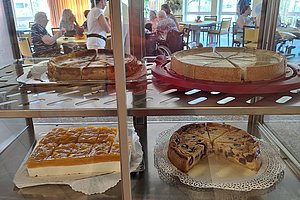 Vier verschiedene Kuchenstücke in einer Glasvitrine in einem Café mit Gästen im Hintergrund