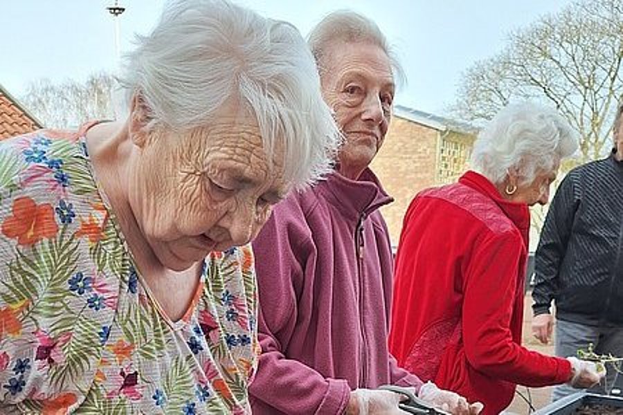 Mehrere ältere Personen stehen an einem Hochbeet und arbeiten mit Erde, eine trägt ein buntes Blumenmusterhemd, eine andere eine rote Jacke.
