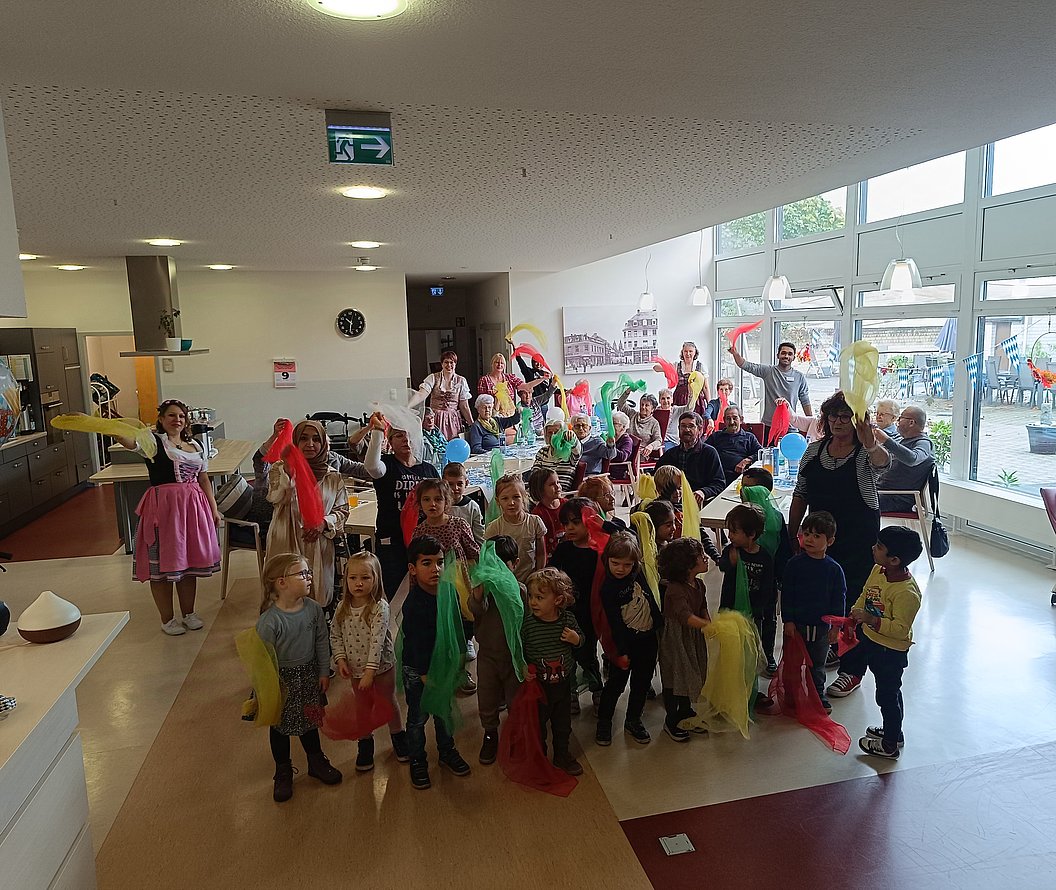 Gruppe von Kindern und Erwachsenen in einem hellen Raum, viele halten bunte Tücher, einige Erwachsene tragen traditionelle Trachten.