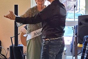 Zwei Personen stehen in einem Raum mit großen Fenstern, eine Person trägt eine schwarze Jacke und Jeans, die andere ein helles Kleid.