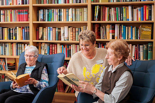 Drei ältere Frauen sitzen vor einem Bücherregal, zwei lesen gemeinsam ein Buch, die dritte liest allein.