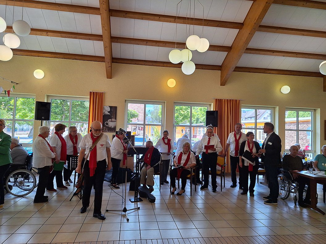 Gruppe älterer Menschen in einem Saal mit Holzbalkendecke, einige sitzen, andere stehen, mehrere Mikrofone und Fenster im Hintergrund.